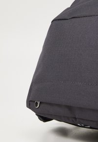 Angolo di una borsa di tessuto grigia con una superficie texturizzata, caratterizzata da una cucitura visibile e un anello metallico. Il design è minimalista e compatto.