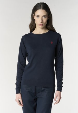 Giovane donna con lunghi capelli scuri che indossa un maglione a maniche lunghe blu navy con un piccolo emblema rosso sul petto.