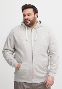 Mann i lys grå zip-up hettejakke med hender i lommene, ser svakt nedover og til venstre, står mot en ensfarget hvit bakgrunn.