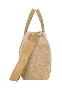 Sac beige texturé avec un extérieur doux et duveteux, deux courtes poignées et une base large et plate. Doté d'une bandoulière en toile avec des lettres blanches.