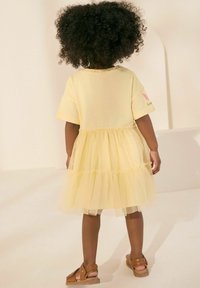 Robe jaune en coton avec une jupe en tulle, dotée de manches courtes et d'un design à volants superposés. Des sandales brunes complètent le look.