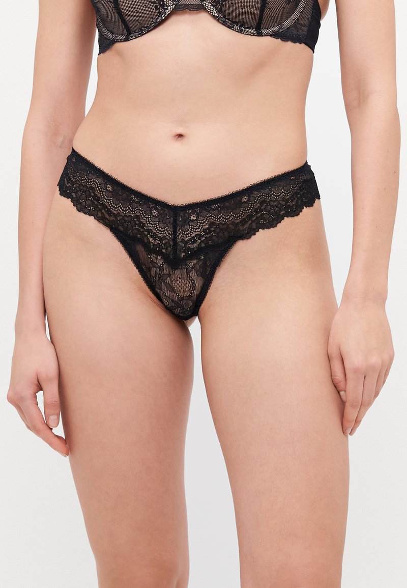 Black lace thong featuring delicate floral patterns, a scalloped waistband, and sheer craftsmanship. Thin sides enhance minimal coverage.