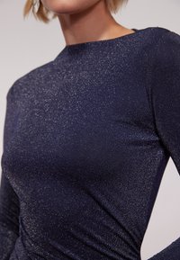Top a maniche lunghe blu navy realizzato in tessuto scintillante con una consistenza liscia e scollatura a barca, caratterizzato da una silhouette aderente e un delicato dettaglio drappeggiato.