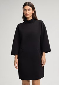 Zwarte jurk met een hoge hals, brede mouwen en zijzakken. Gemaakt van een gladde stof, met een ontspannen silhoette en een knie-lengte snit.