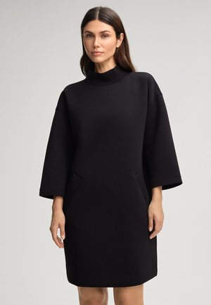 Zwarte jurk met een hoge hals, brede mouwen en zijzakken. Gemaakt van een gladde stof, met een ontspannen silhoette en een knie-lengte snit.