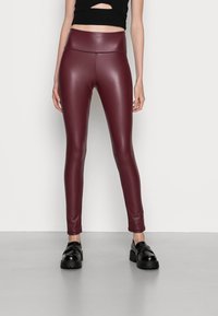 Burgundowe legginsy z ekoskóry z wysokim stanem, zwężanym krojem, gładką fakturą i błyszczącym wykończeniem, w połączeniu z czarnymi butami na platformie.