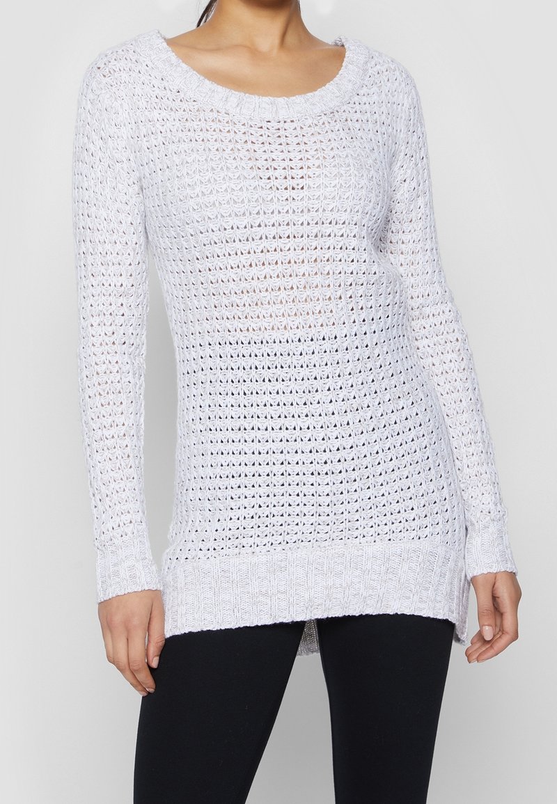 Femme portant un pull en maille blanc à manches longues, à col rond et motif texturé en maille ajourée, associé à un pantalon noir.