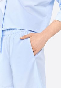 Chemise légère à manches courtes bleu clair avec un col, dotée d'une taille élastique et d'une poche latérale ; tissu lisse et léger.