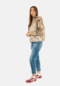 Giacca con cappuccio in oro metallico con polsini elasticizzati, abbinata a jeans skinny blu e sneaker rosse con dettagli bianchi.