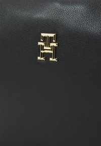 Borsa a tracolla in pelle nera con un emblema dorato con lettere intrecciate. La superficie presenta una finitura testurizzata e bordi lisci.