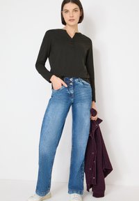 Dunkles, langärmliges Henley-Oberteil, mittelblaue Jeans mit weitem Bein und fünf Taschen, Knopfverschluss, dazu eine bordeauxrote Fleecejacke über dem Arm gelegt.