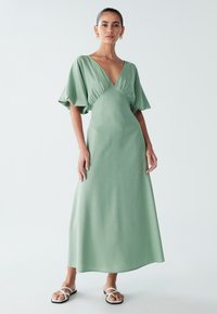 Lång, figurnära klänning i ett mjukt grönt tyg, med djup V-ringning, korta puffärmar och en slät, odekorerad yta.