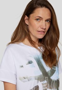 Weißes Baumwoll-T-Shirt in lockerer Passform mit einem grafischen Design in Blau- und Grüntönen, ergänzt durch texturierte Akzente und einem runden Halsausschnitt.