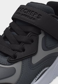 Gros plan sur une chaussure de sport Skechers noire et grise avec des lacets texturés, une languette rembourrée et une bride marquée sur le dessus.