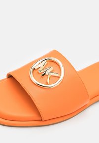 Sandale à glissière orange avec une large bride ornée d'un emblème circulaire doré et une semelle plate sur fond blanc.