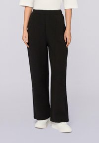 Persoon in zwarte wijde broek met elastische taille en witte sneakers, staand tegen een effen lichte achtergrond.