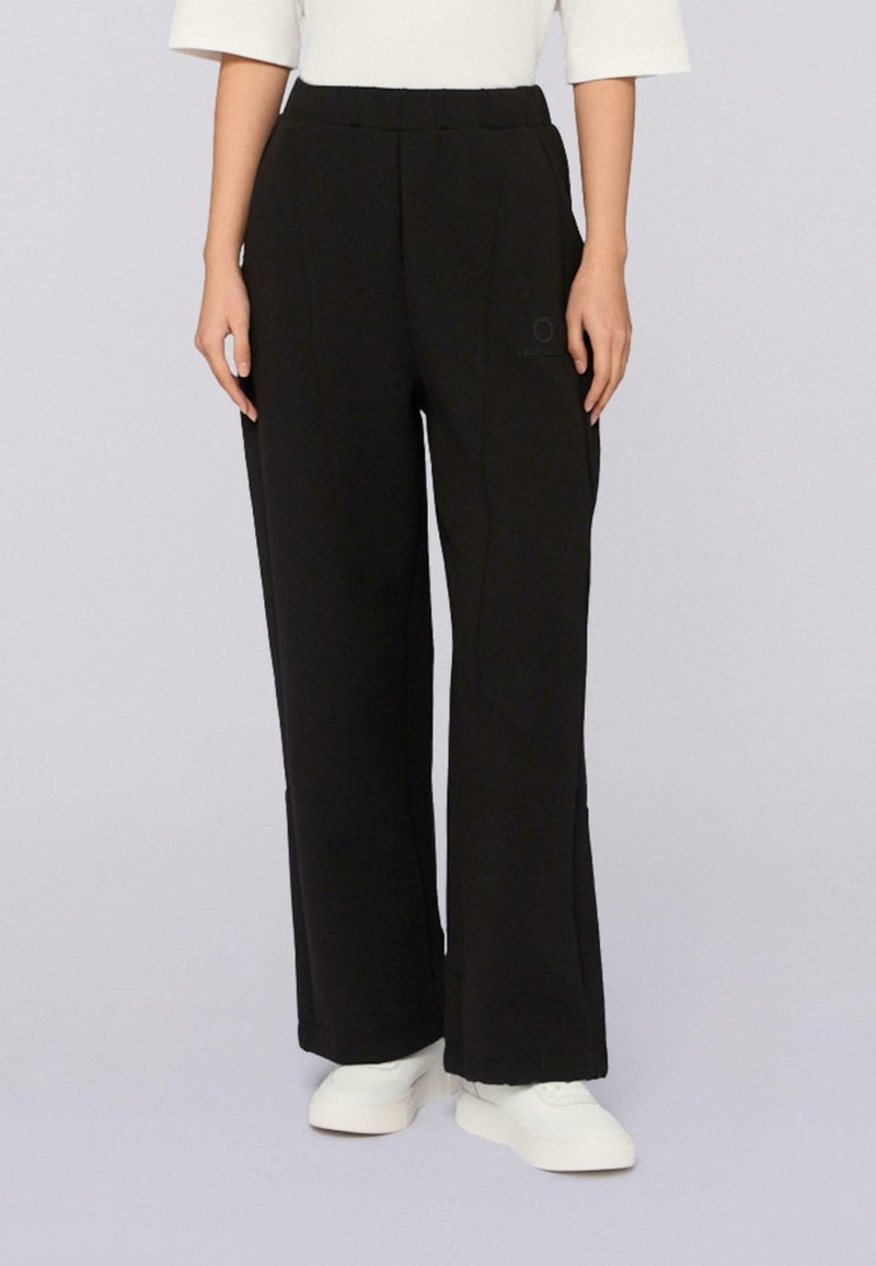 Persoon in zwarte wijde broek met elastische taille en witte sneakers, staand tegen een effen lichte achtergrond.