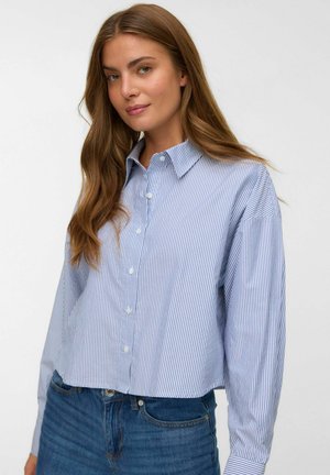 Woman with long brown hair wearing a blue and white vertical striped button-up shirt and blue jeans, standing against a white background.