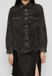 Giacca di jeans nera con una texture sbiadita, chiusura a bottoni, due tasche sul petto e colletto. Indossata su una gonna nera con uno spacco laterale.