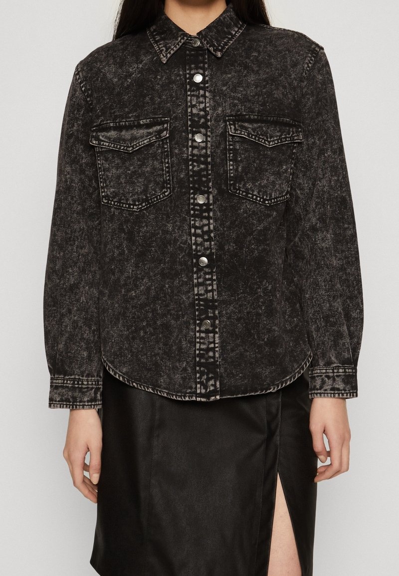 Giacca di jeans nera con una texture sbiadita, chiusura a bottoni, due tasche sul petto e colletto. Indossata su una gonna nera con uno spacco laterale.
