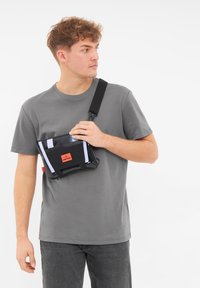 Svart axelremsväska med vita remmar, röd etikett och blanka detaljer. Visad på en person som bär en grå tröja och mörka jeans.