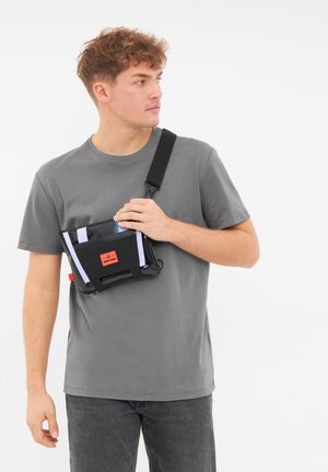 Borsa a tracolla nera con cinghie bianche, etichetta rossa e dettagli lucidi. Presentata su una persona che indossa una maglietta grigia e jeans scuri.