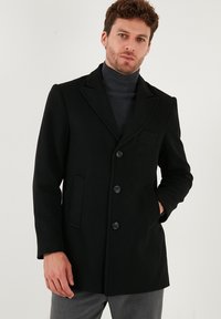 Melna vilnas mēteles ar iegriezumu apkakli, trim pogām un divām sānu kabatām. Valkāta virs tumša turtlenecka ar teksturētu un pieguļošu dizainu.