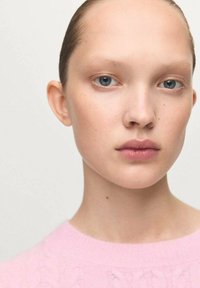 Primo piano di un volto con pelle liscia, occhi azzurro chiaro e lentiggini sottili. Il modello indossa un maglione rosa a trecce. I capelli sono raccolti all'indietro.