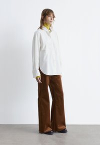 White button-up shirt with rounded hem over a bright yellow top, paired with wide brown corduroy pants and black shoes, against a simple background.