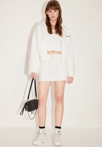 Sudadera corta blanca, top blanco sin mangas y falda mini a juego. Los accesorios incluyen un bolso acolchado negro y zapatillas blancas de suela gruesa.