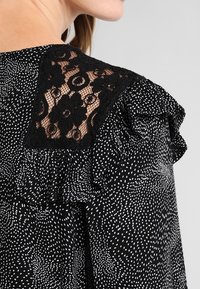 Blusa nera con pois bianchi e dettagli in pizzo sulla spalla. I bordi arricciati aggiungono texture al design.