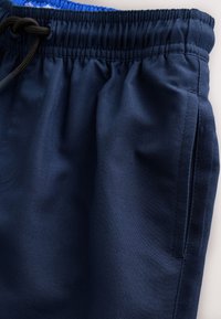 Tmavomodré športové šortky z ľahkej tkaniny. Majú elastický pás so sťahovacou šnúrkou a bočné vrecká. Hladká štruktúra.