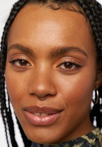 Närbild av en person med flätat hår, en guldfärgad septumring och naturlig sminkning med varma toner och glansiga, klara läppar.