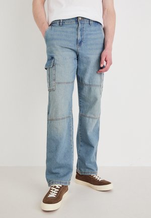 Osoba w jasnoniebieskich cargo spodniach z denimu z bocznymi kieszeniami i brązowych trampkach z białymi sznurowadłami stojąca na jednolitym tle.