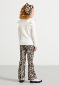 Pull blanc avec des accents de volants aux épaules, associé à un pantalon évasé à motifs léopard et des chaussures argentées. La coiffure de l'enfant comporte un grand nœud léopard.