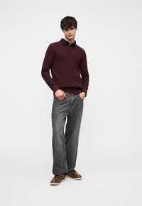 Pull en tricot marron avec un col bleu marine, associé à un jean gris jambes larges et des chaussures marron. Comprend des poignets côtelés et un détail logo subtil.