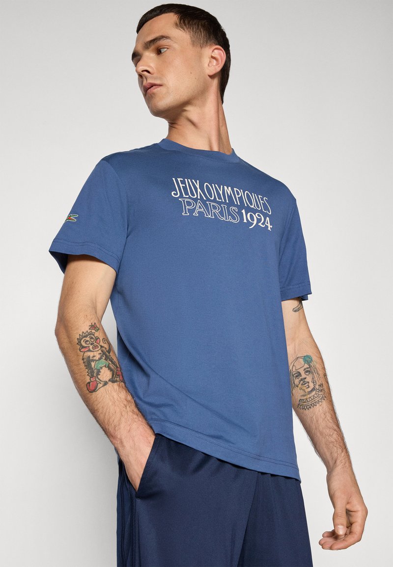 T-shirt en coton bleu avec "JEUX OLYMPIQUES PARIS 1924" imprimé en blanc. Manches courtes et col rond. Porté avec un short bleu marine.