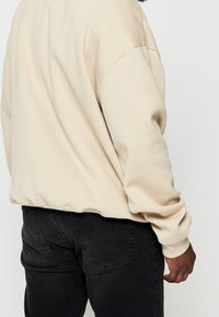 Sweat-shirt beige à coupe décontractée avec des poignets côtelés, associé à un jean noir foncé avec poches arrière renforcées. Texture lisse.