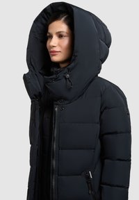 Kvinde iført en sort dunjakke med en stor polstret hætte, lynet op, vendt mod venstre mod en ensfarvet grå baggrund.