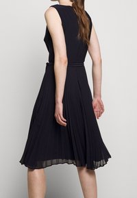 Žena s dlouhými vlasy, oblečená v bezramínkových černých plisovaných šatech po kolena s tenkým opaskem, zobrazená zezadu na jednolitém pozadí.