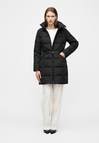 Γυναίκα φορώντας μαύρο μακρύ παλτό με επένδυση και ψηλό γιακά, λευκά παντελόνια και μαύρα μυτερά παπούτσια, όρθια με το ένα χέρι στην τσέπη του παλτού.