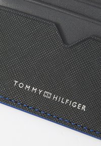 Svart teksturert lommebok med hevet logo, blå sømmer og flere kortlommer. Designet med en slank form og holdbare materialer.