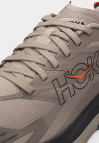 Κοντινή όψη μπεζ παπουτσιού τρεξίματος Hoka με υφή υφάσματος, μπεζ κορδόνια και μαύρη σόλα, με πορτοκαλί και μαύρο λογότυπο Hoka στο πλάι.