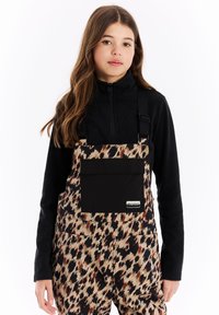 Salopette stampata con un motivo leopardato colorato, dettagli neri, spalline regolabili e una tasca frontale; indossata su una maglietta nera a coste.