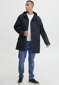 Marine vanntett jakke med hette, med knappelukking, en frontlomme og en avslappet passform. Brukt over en hvit skjorte og sammen med blå jeans.
