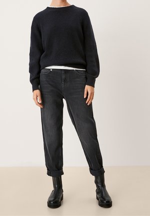 Osoba ubrana w czarny sweter dzianinowy, biały podkoszulek, podwinięte ciemnoszare dżinsy i czarne skórzane botki do kostek stoi na jednolitym tle.