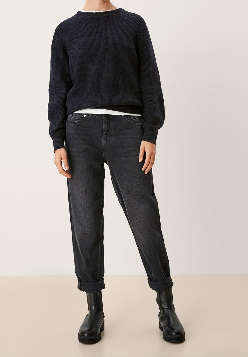 Osoba ubrana w czarny sweter dzianinowy, biały podkoszulek, podwinięte ciemnoszare dżinsy i czarne skórzane botki do kostek stoi na jednolitym tle.