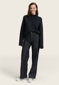 Maglione rigato blu con colletto alto, abbinato a pantaloni larghi in denim scuri e sneakers bianche. Ambientato su uno sfondo beige chiaro.