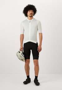 Maillot de cyclisme léger à manches courtes en blanc avec une fermeture éclair à l'avant et un short de cyclisme noir. Le mannequin tient un casque gris avec des accents noirs.