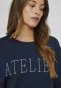 Jūras zila treniņu krekls ar baltiem izšūtiem burtiem "ATELIER", apaļa kakla izgriezums, mīksta tekstūra, ikdienas piegriezums un nedaudz nolaištas plecu šuves.